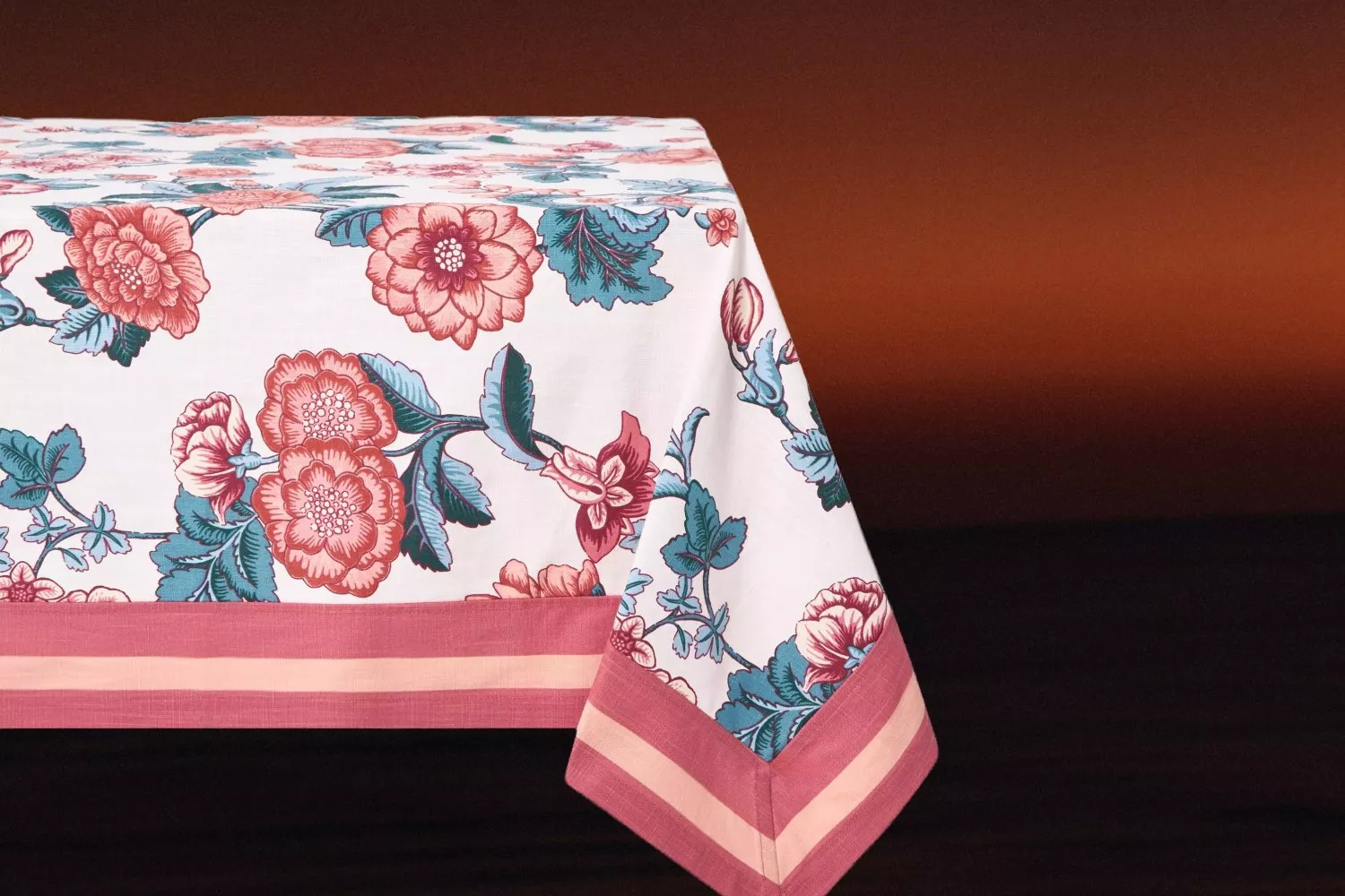 Floral tablecloth with pink border, vintage rose print in teal and coral, draped over dining table corner.