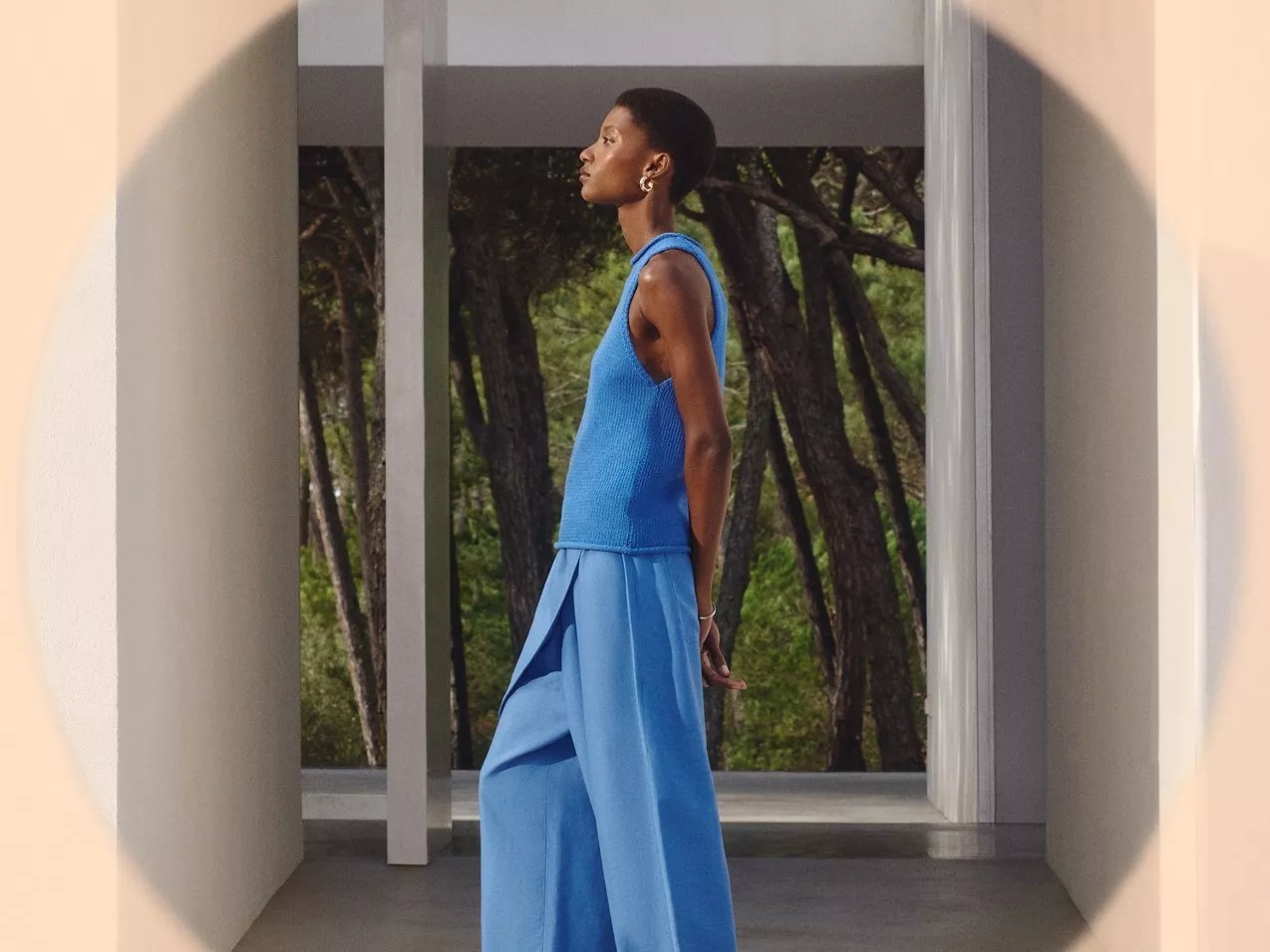 A woman in a blue sleeveless top and trousers, standing in a modern architectural space with trees visible.