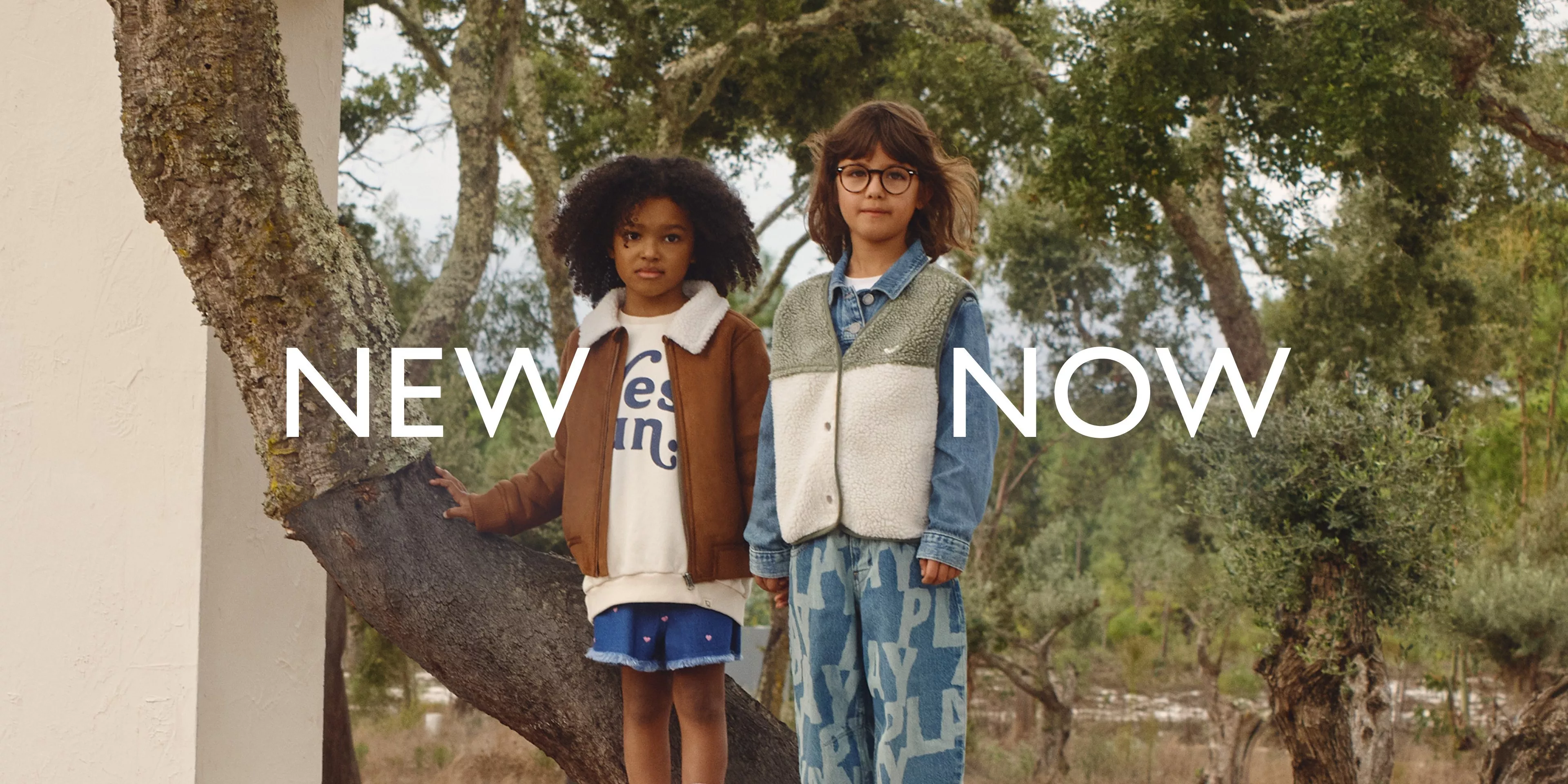 Two children in autumn clothing stand outdoors among trees, with the text "NEW NOW" overlaid.