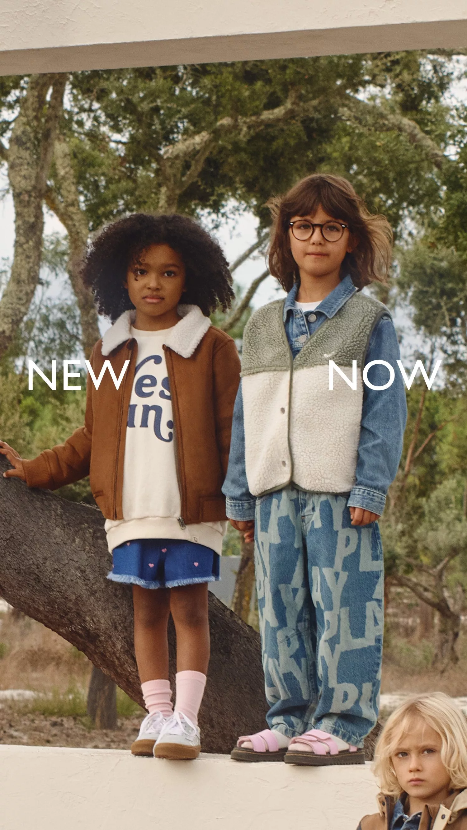Two children in autumn clothing stand outdoors among trees, with the text "NEW NOW" overlaid.