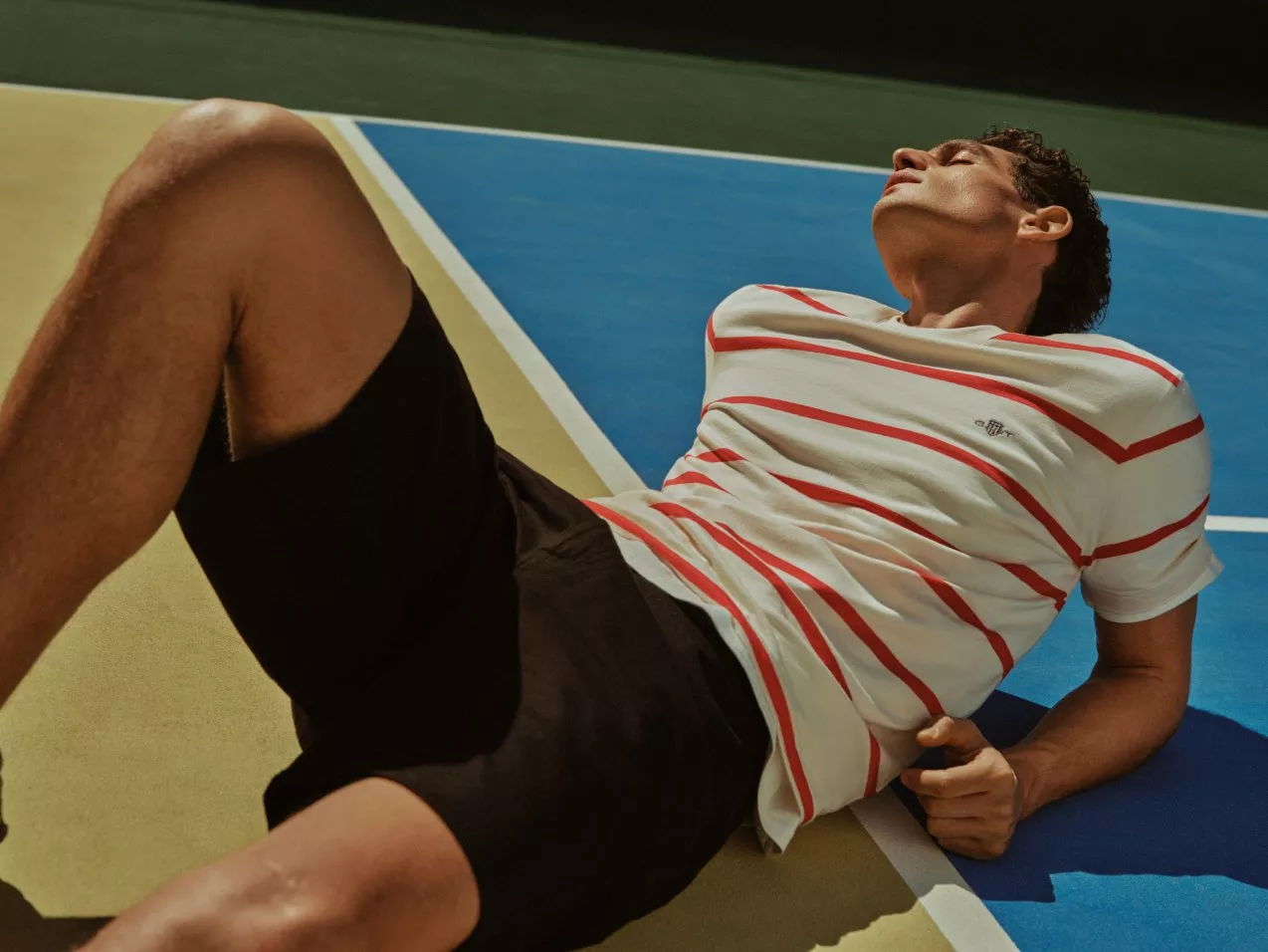 Man lying on outdoor court after match, wearing striped shirt and black shorts in sunlight