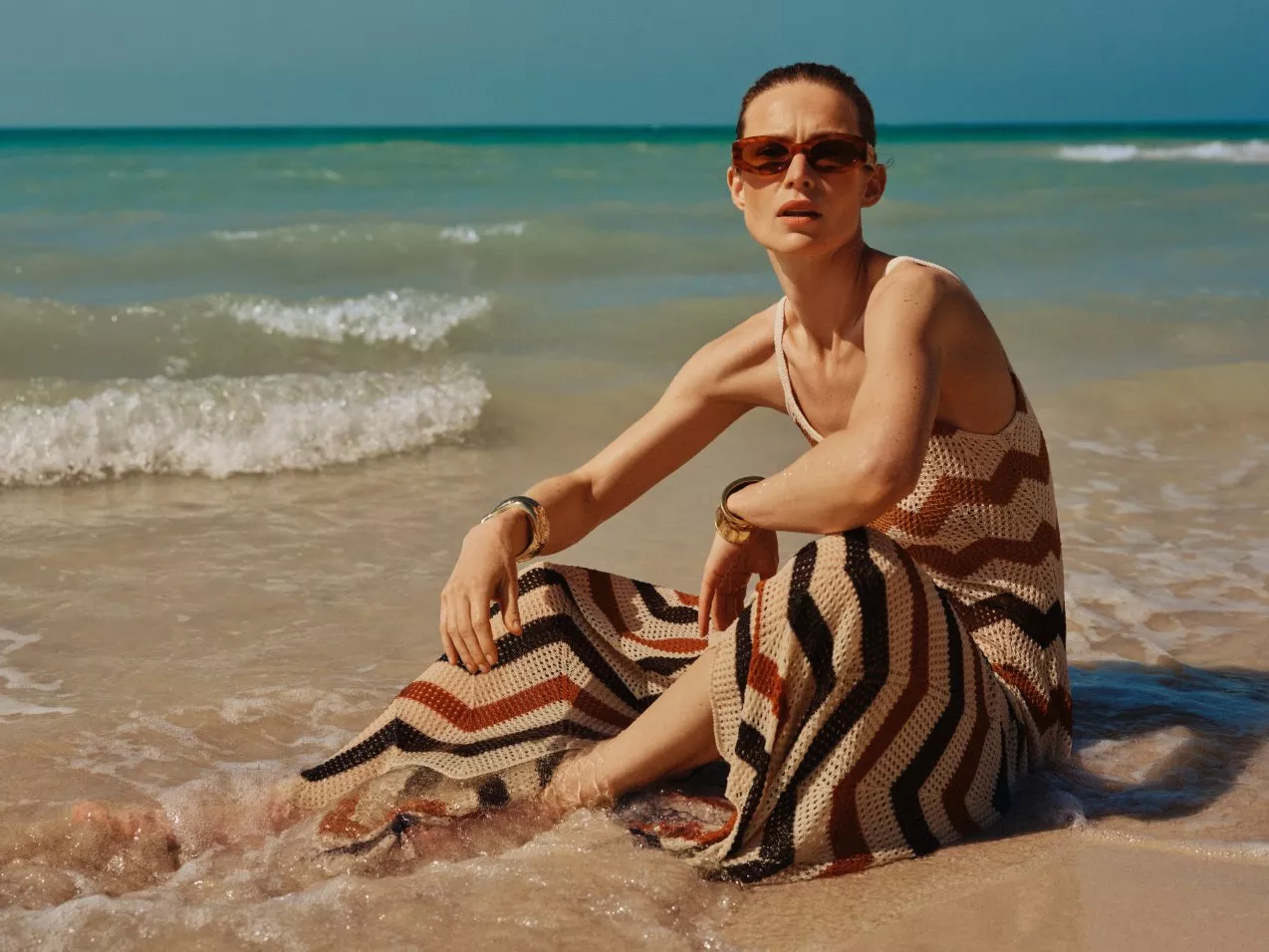 Woman in sunglasses wearing striped crochet maxi dress, seated at shoreline with waves on a sunny beach