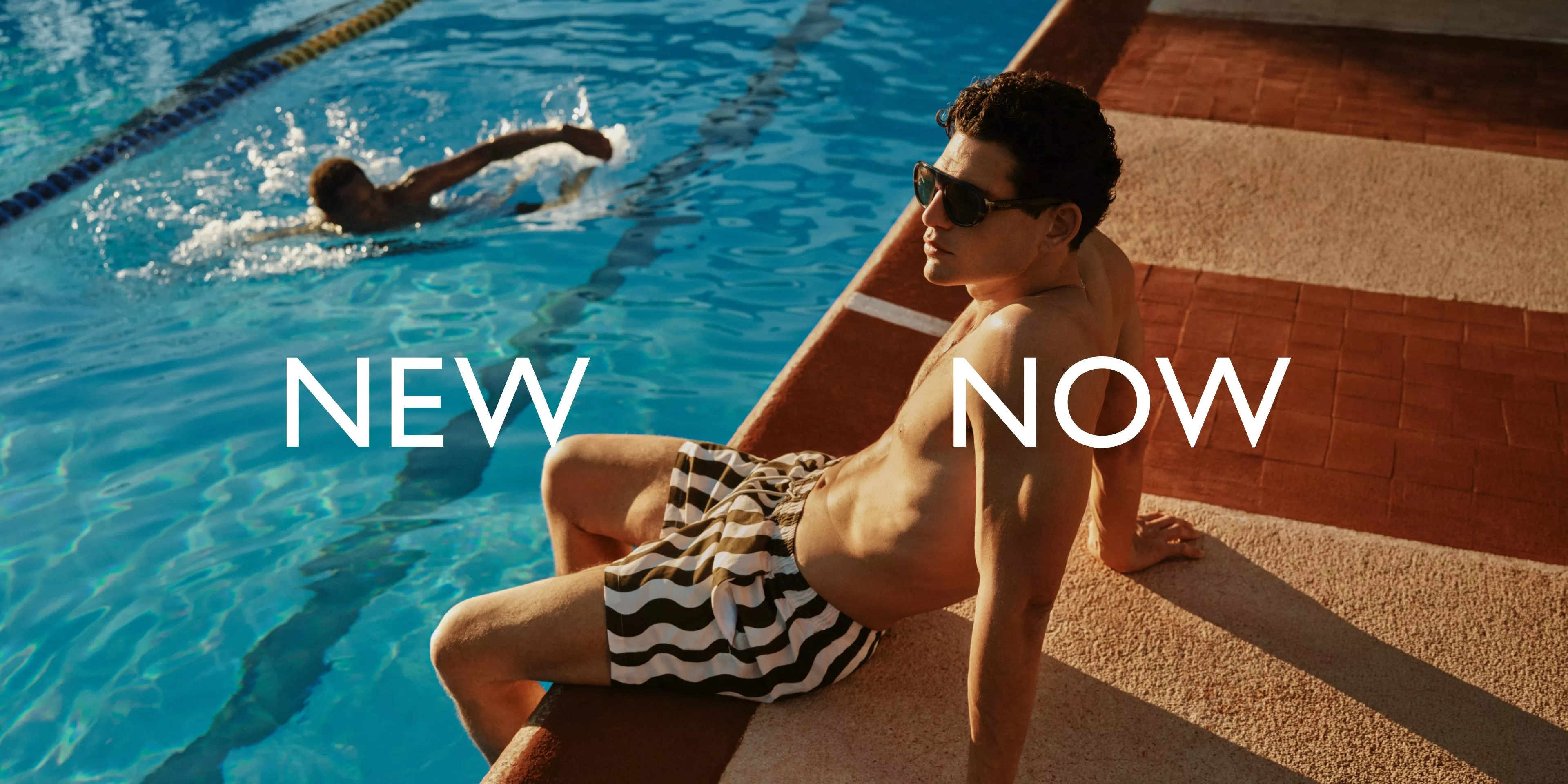 man sitting on the edge of a sunlight pool wearing striped swim shorts and sunglasses