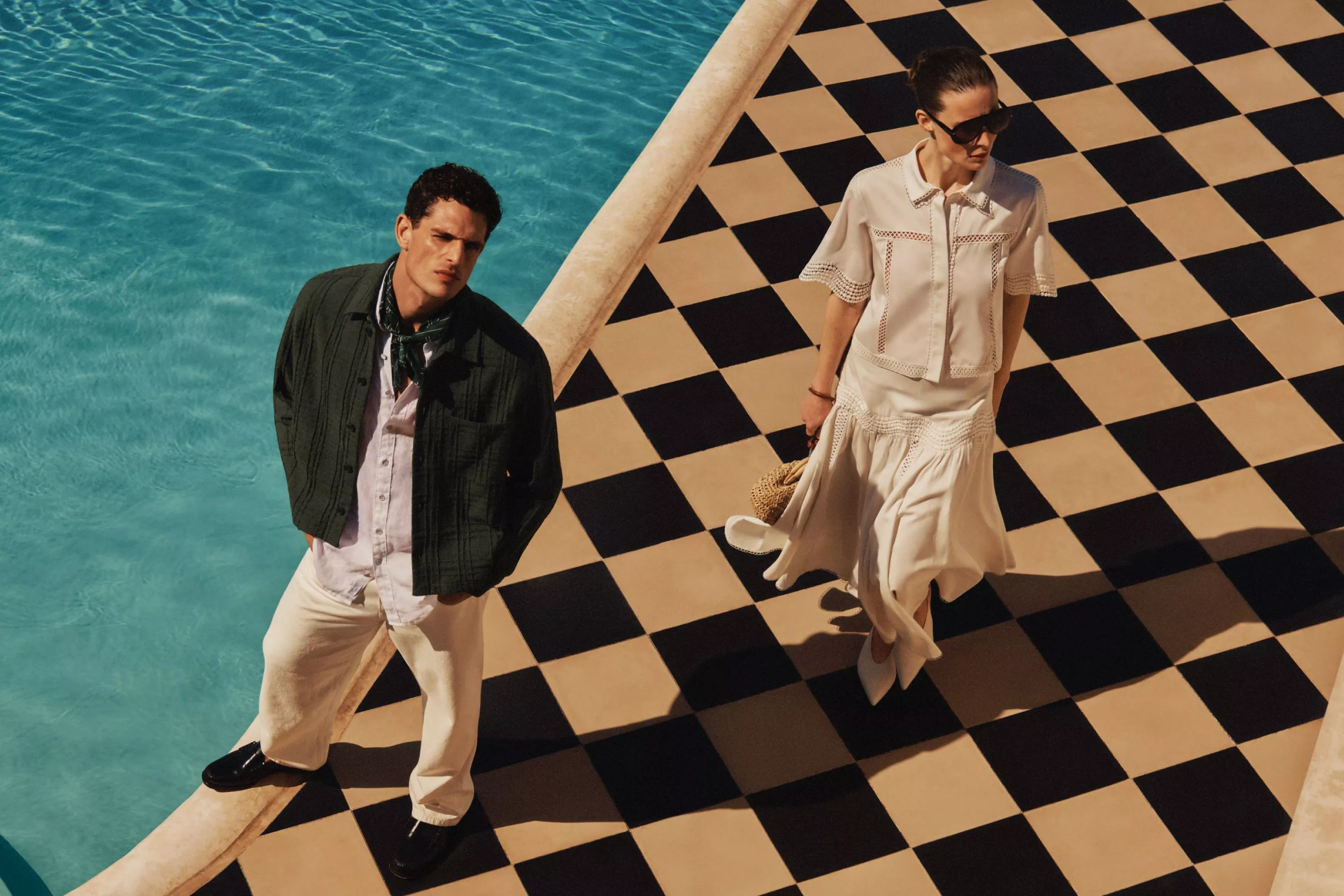 man and women dressed in summer outfits, standing by a pool 