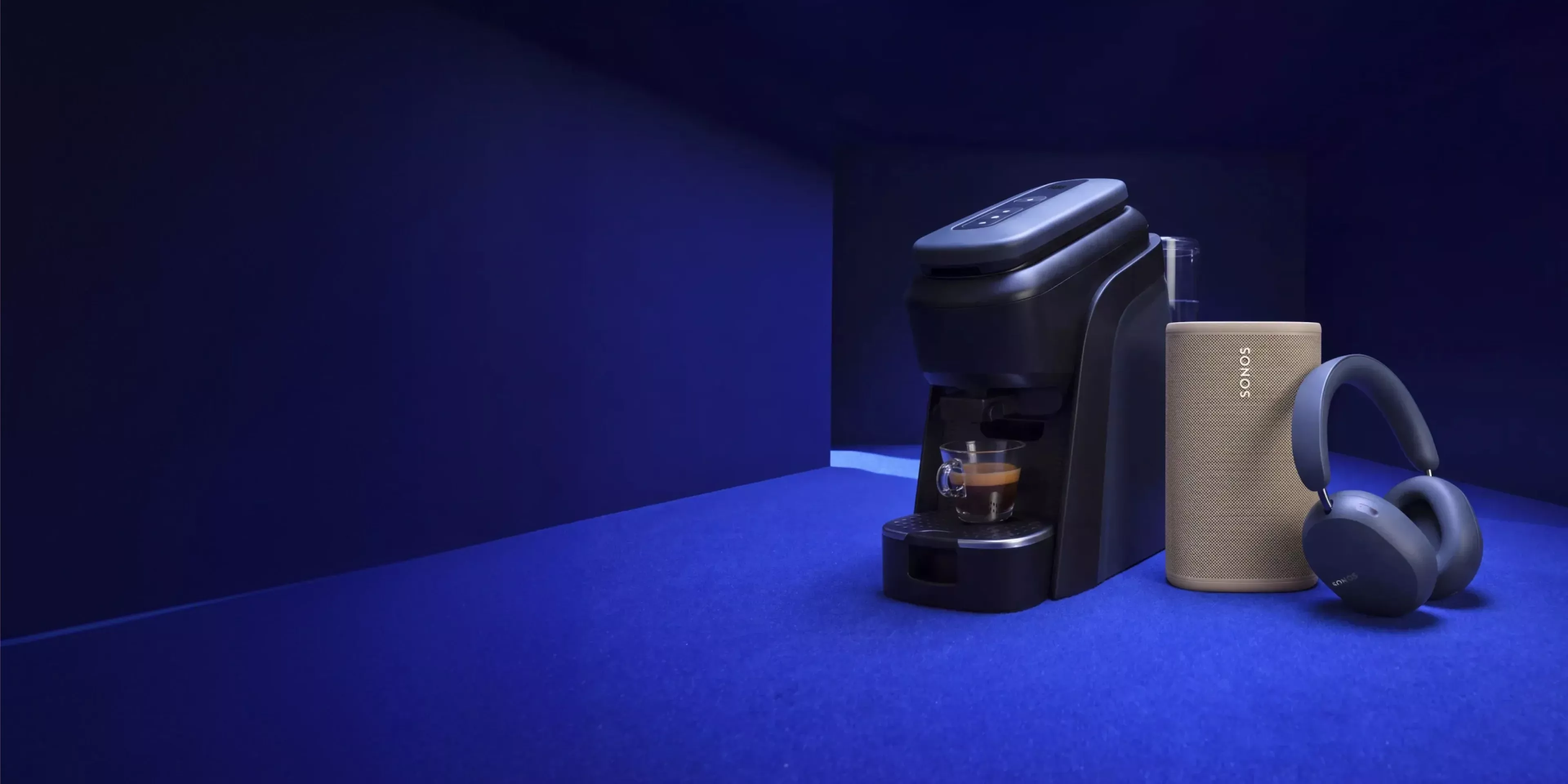Coffee machine brewing espresso beside Sonos speaker and wireless headphones on blue background in studio setup