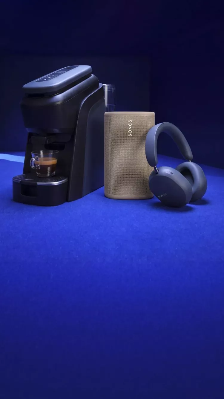 Coffee machine brewing espresso beside Sonos wireless speaker and over-ear headphones on blue surface