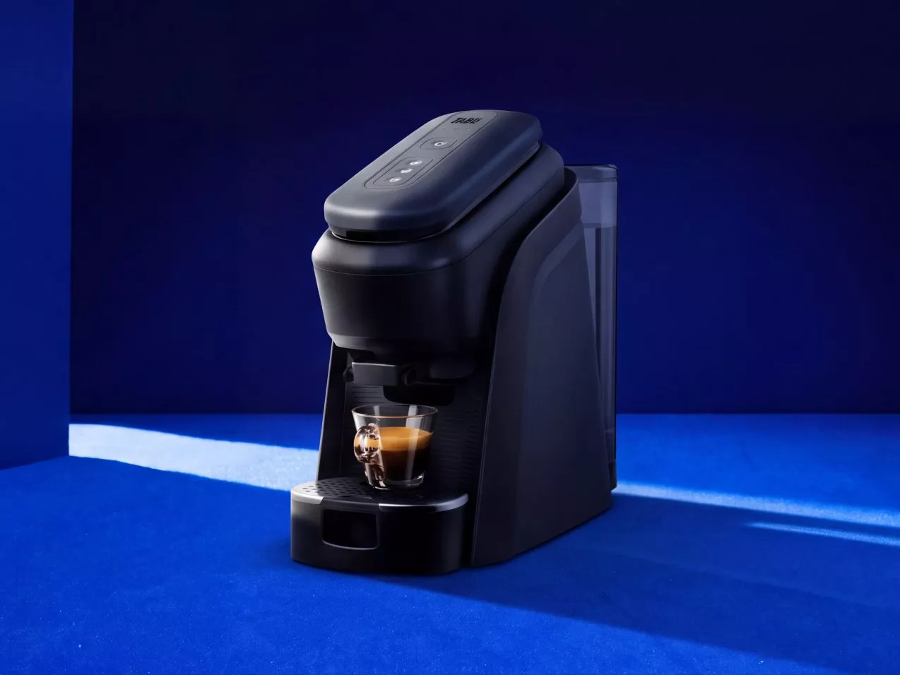 Black pod coffee machine brewing espresso in glass cup on blue background, modern compact countertop appliance