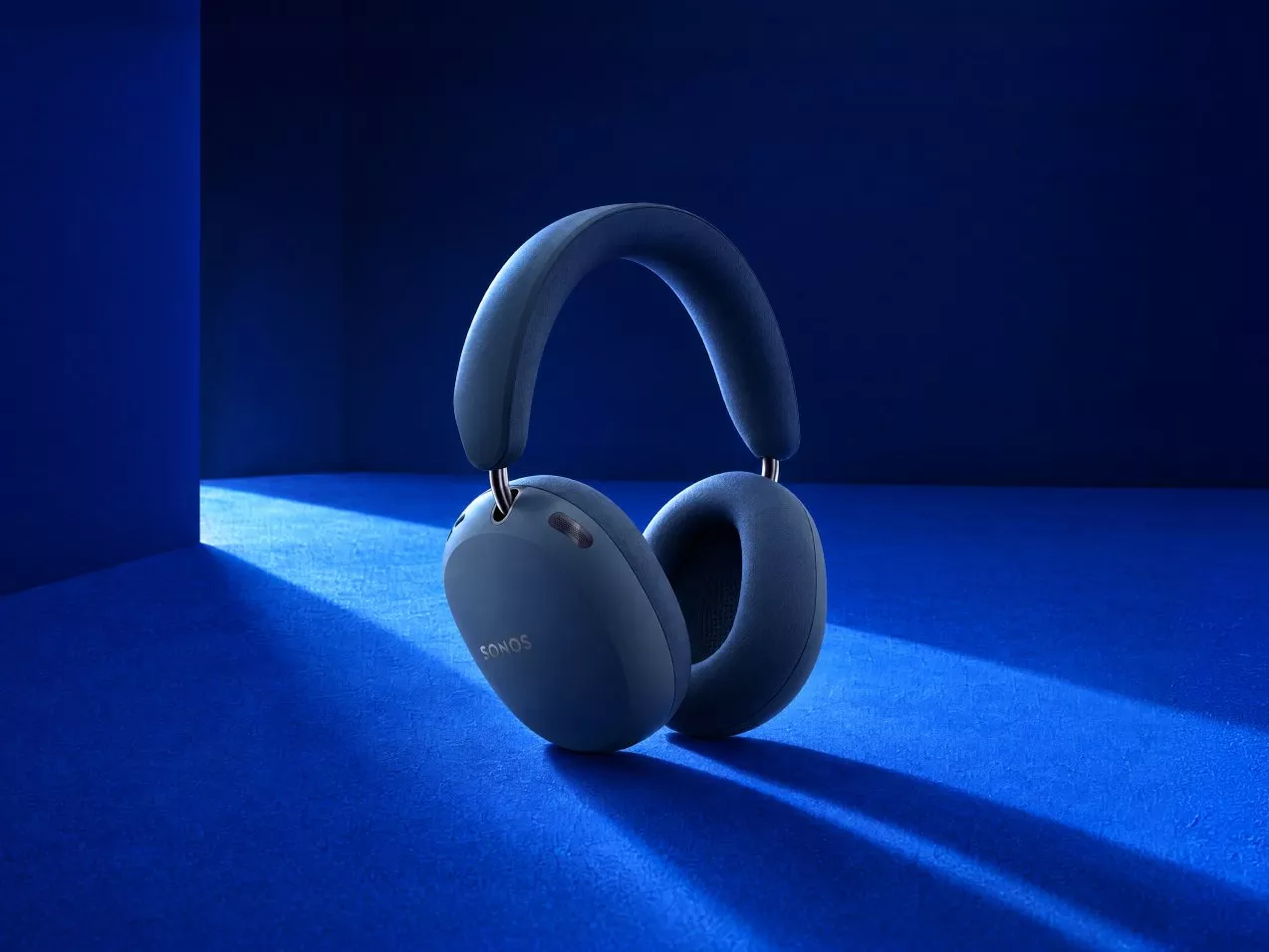 Blue Sonos over-ear wireless headphones on a deep blue studio background with dramatic lighting and shadow