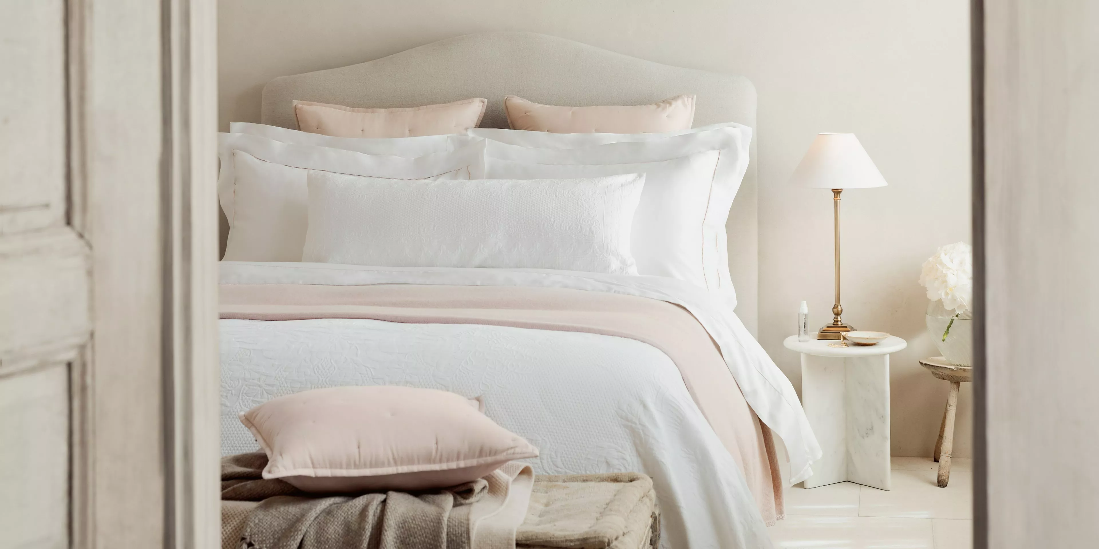 Elegant bedroom with upholstered headboard, white and blush bedding, bedside lamp and marble side table