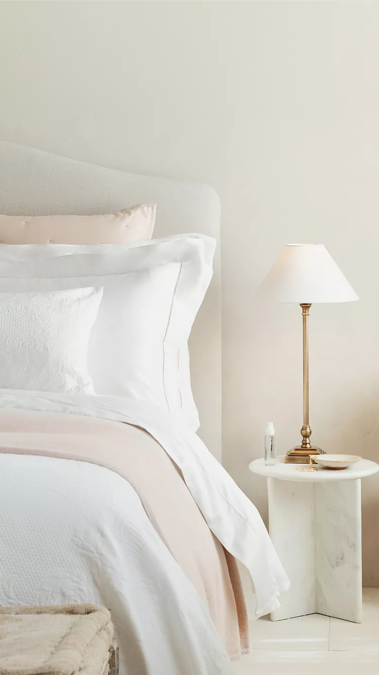 Elegant bedroom with upholstered headboard, white and blush bedding, bedside lamp and marble side table
