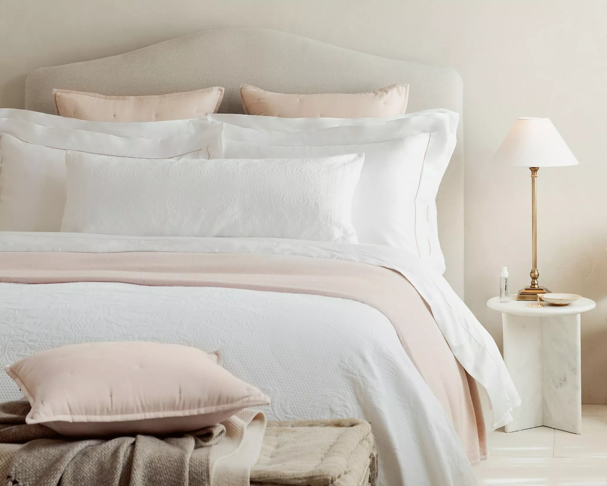 Neutral bedroom with upholstered headboard, white quilted bedding, blush cushions, and bedside lamp on table