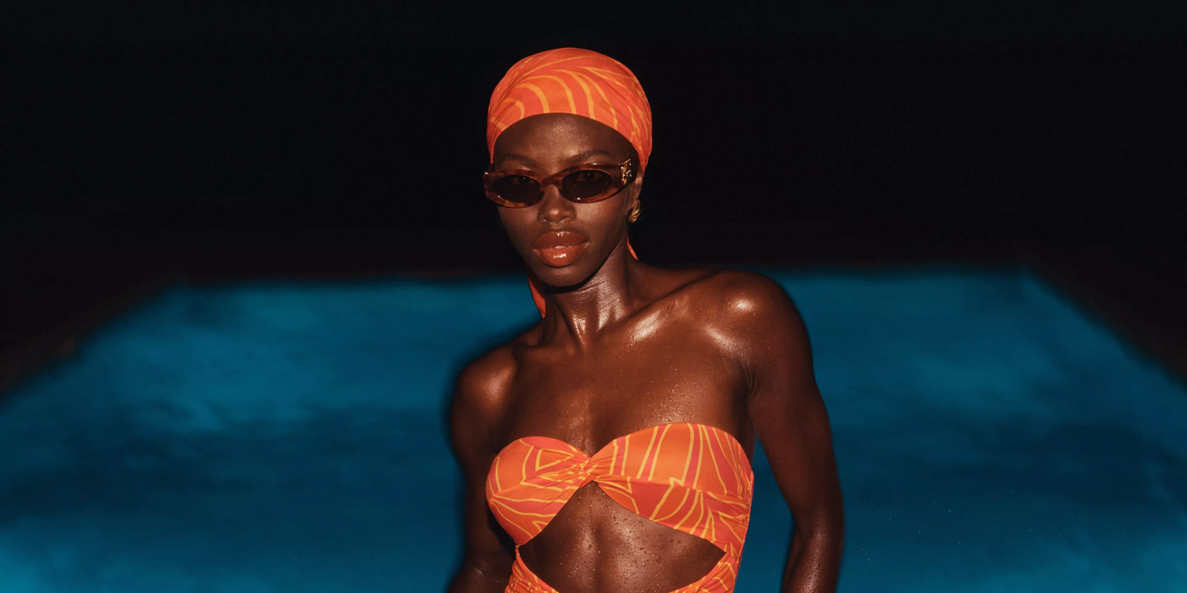 Woman in orange bikini, headscarf and sunglasses by swimming pool at night, glossy skin