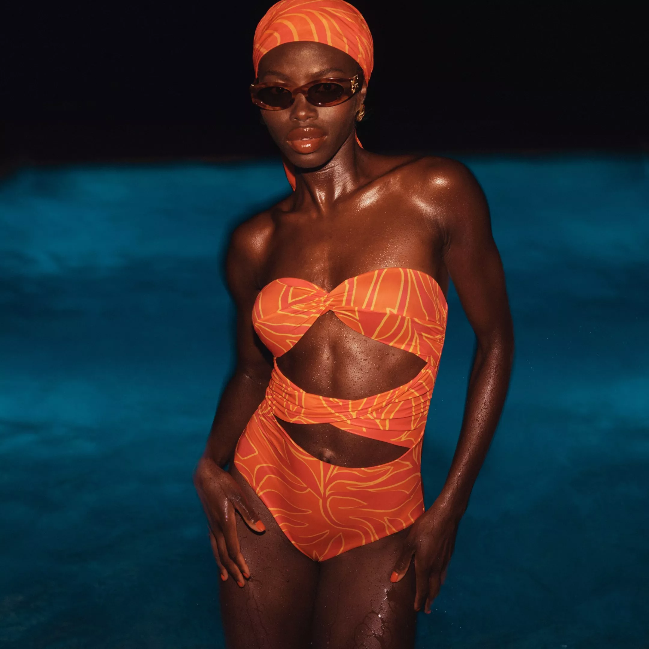 Fashion model in orange cut-out swimsuit and matching headscarf, wearing sunglasses by a blue swimming pool at night