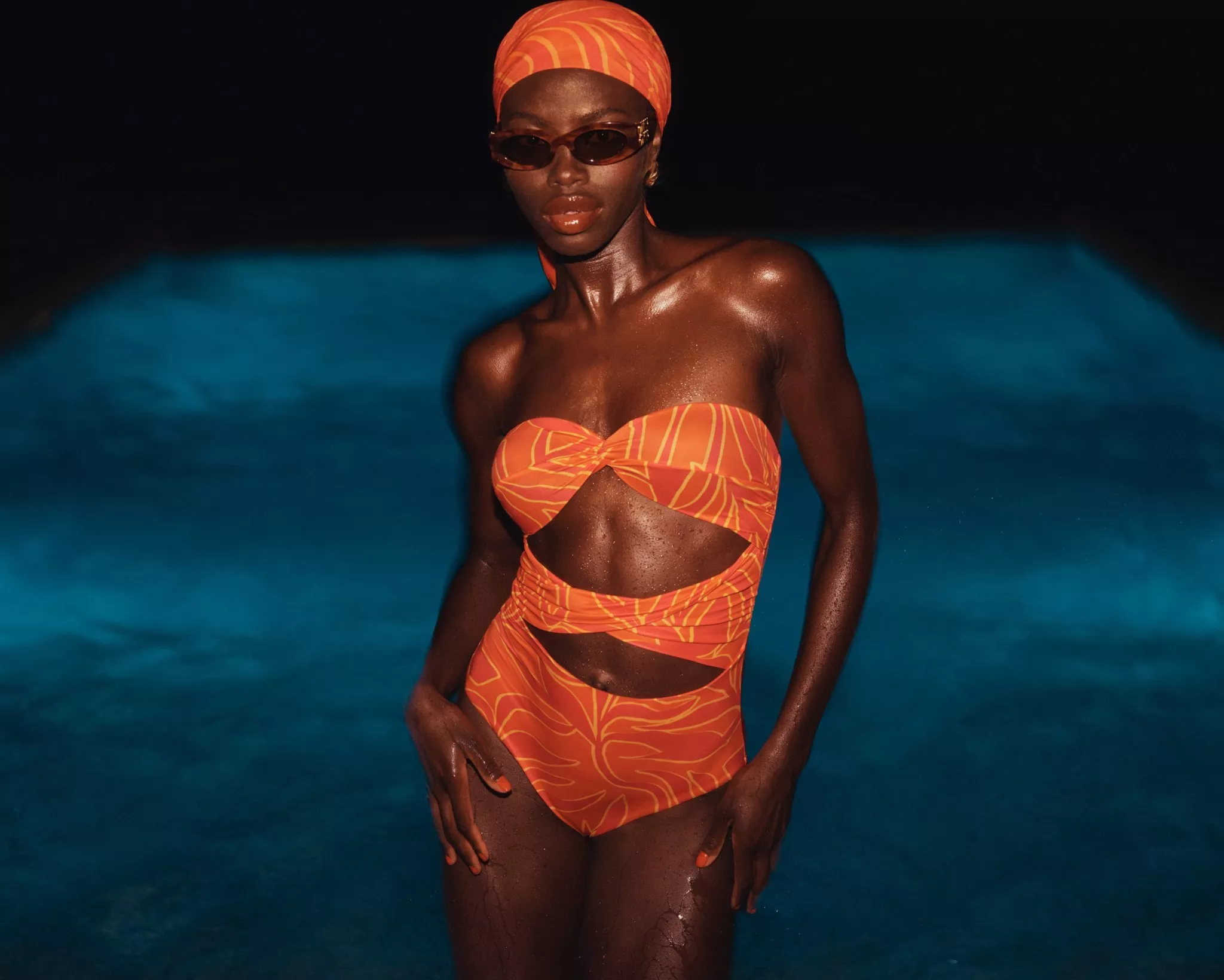 Woman in orange bikini, headscarf and sunglasses by swimming pool at night, glossy skin