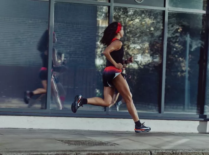 Female runner sprinting past glass building, wearing red headband and black kit, urban fitness training outdoors