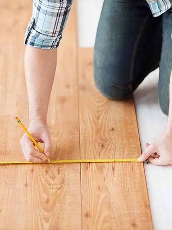 Person measuring wooden floor panels with a tape measure and pencil, wearing a plaid shirt and jeans.