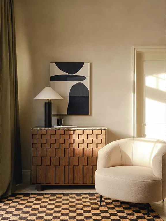 A stylish living room with a textured wooden sideboard, abstract artwork, a cosy chair, and checkered rug.