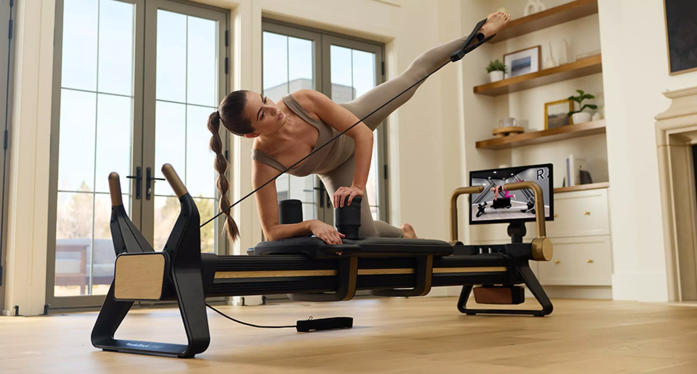 Woman doing Pilates reformer side leg lift with resistance strap in bright home studio workout space