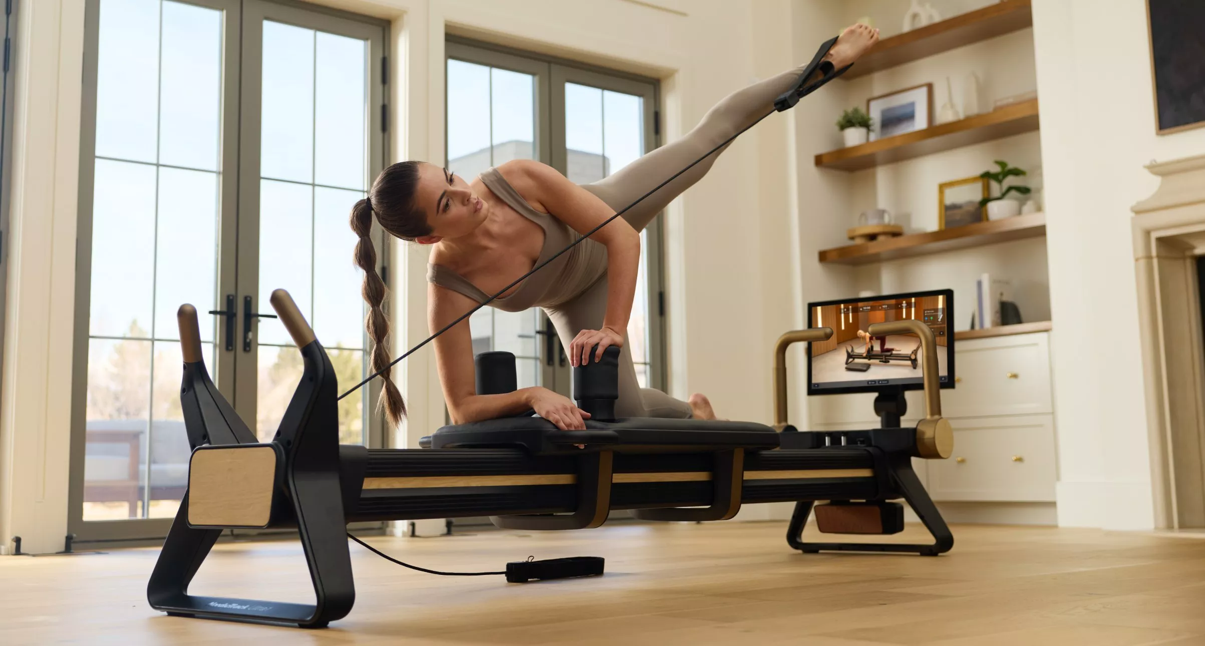 Woman doing Pilates reformer side leg lift with resistance strap in bright home studio workout space
