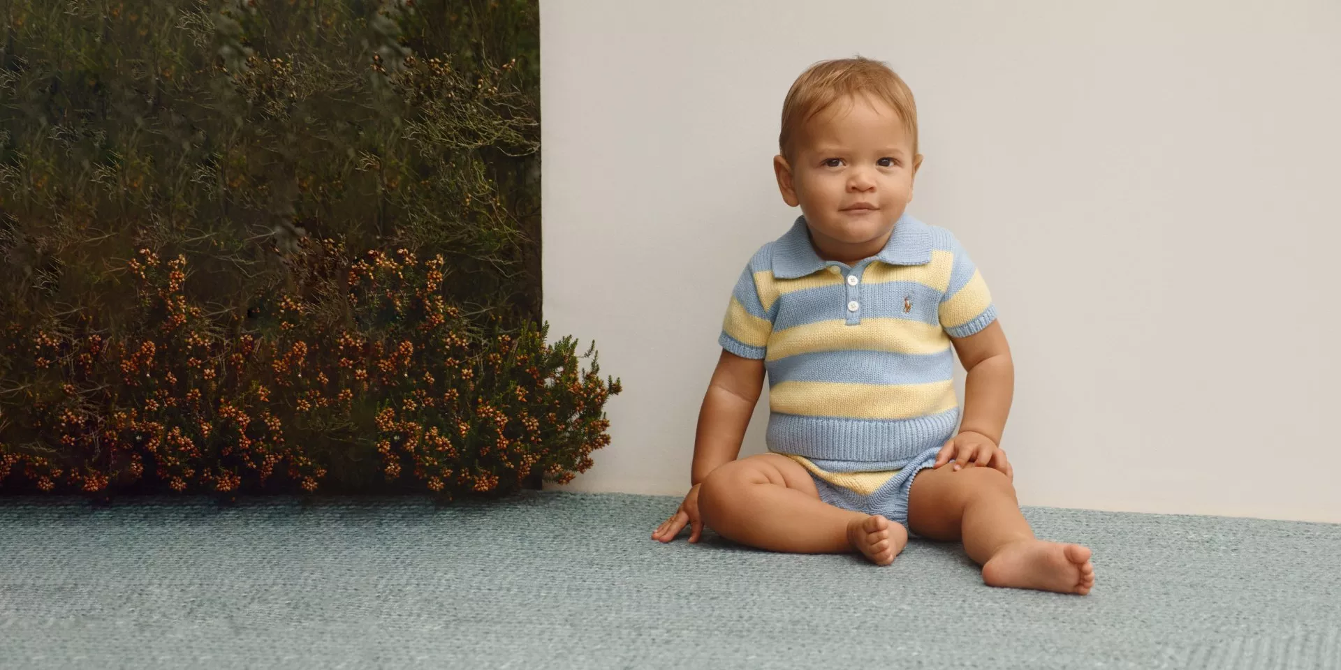 Baby boy sitting on blue rug wearing blue and yellow striped knit romper, studio portrait with neutral backdrop