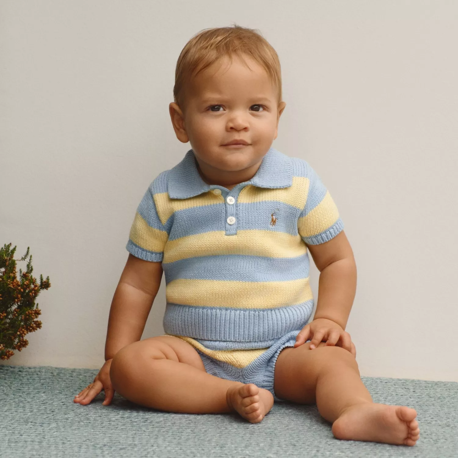 Toddler boy sitting on carpet in blue and yellow striped knit polo romper, studio portrait on neutral background