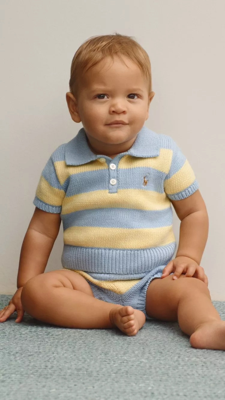 Smiling baby boy in blue and yellow striped knitted romper, sitting on carpet against neutral studio backdrop