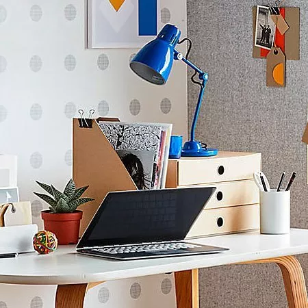 Modern desk with a laptop, blue lamp, potted plant, and organisers. Polka dot wallpaper in background.