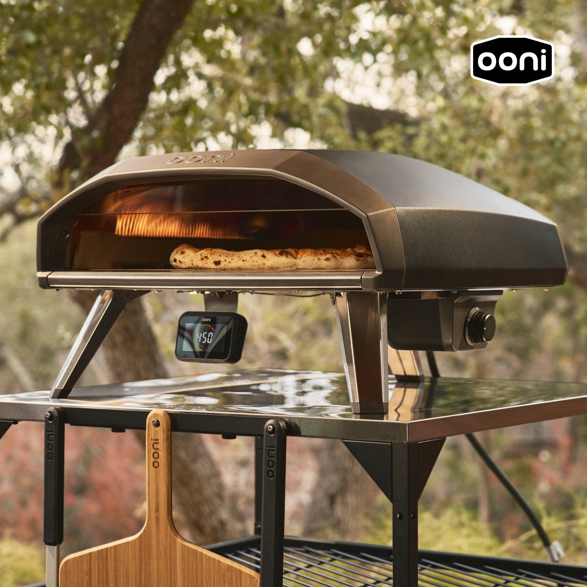 Pizza baking in an outdoor pizza oven with trees in the background.