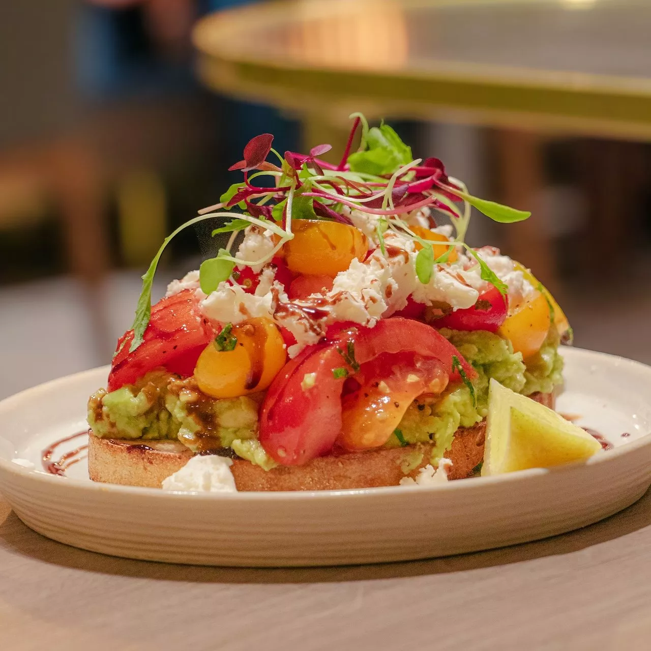 A plate of vibrant avocado toast topped with tomatoes, crumbled cheese, microgreens, and a lemon wedge garnish.
