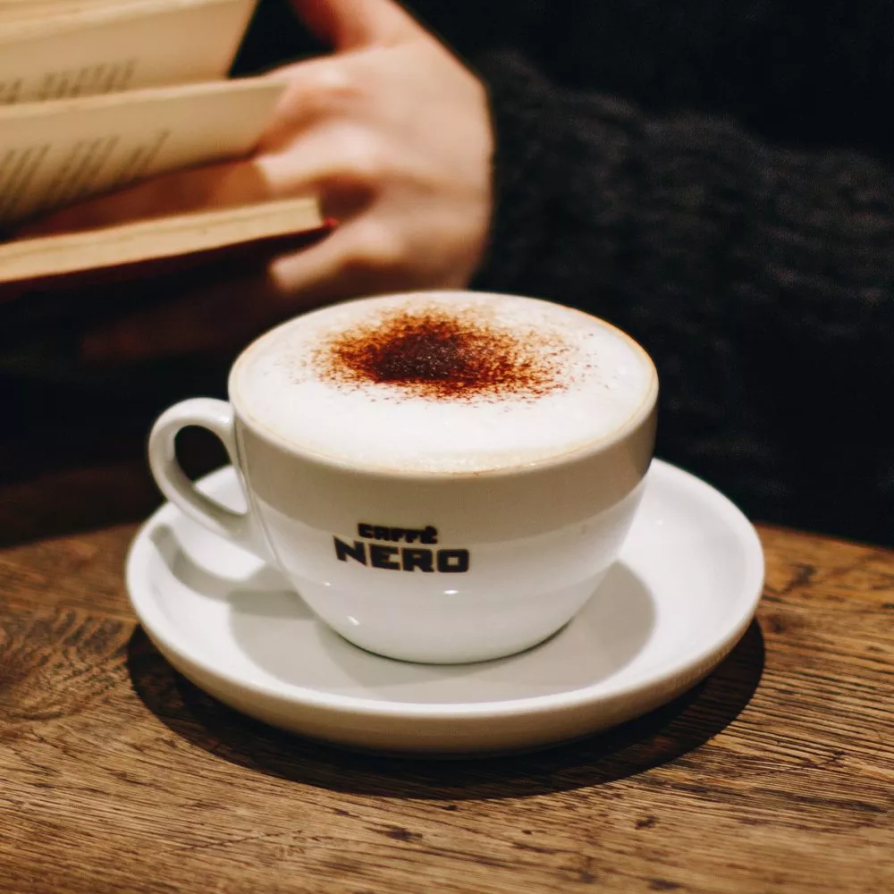 A cappuccino with chocolate topping in a Caffè Nero cup on a wooden table, with a person reading a book nearby.