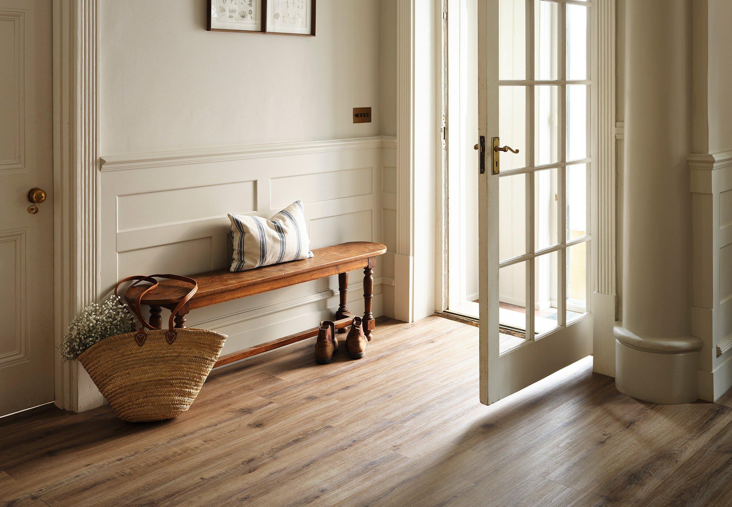 Luxury vinyl hallway with white walls and white door. Wooden bench with a bag next to it. Stripey pillow sat on top of the bench. Frames hung on the wall.