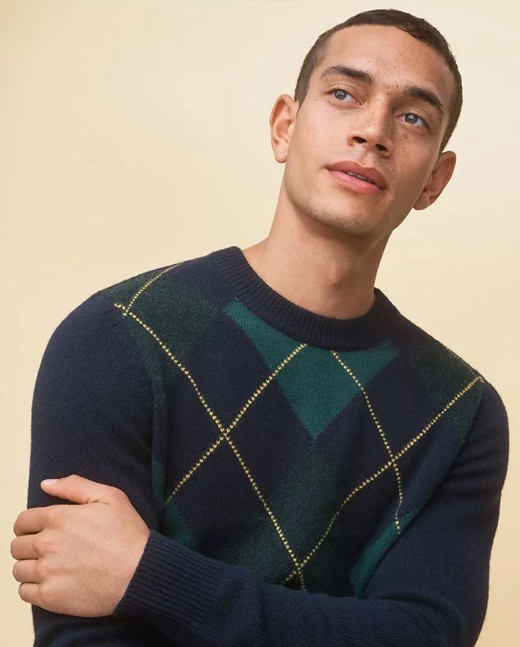 Man wearing a navy and green argyle sweater, looking upwards with a neutral expression against a beige background.