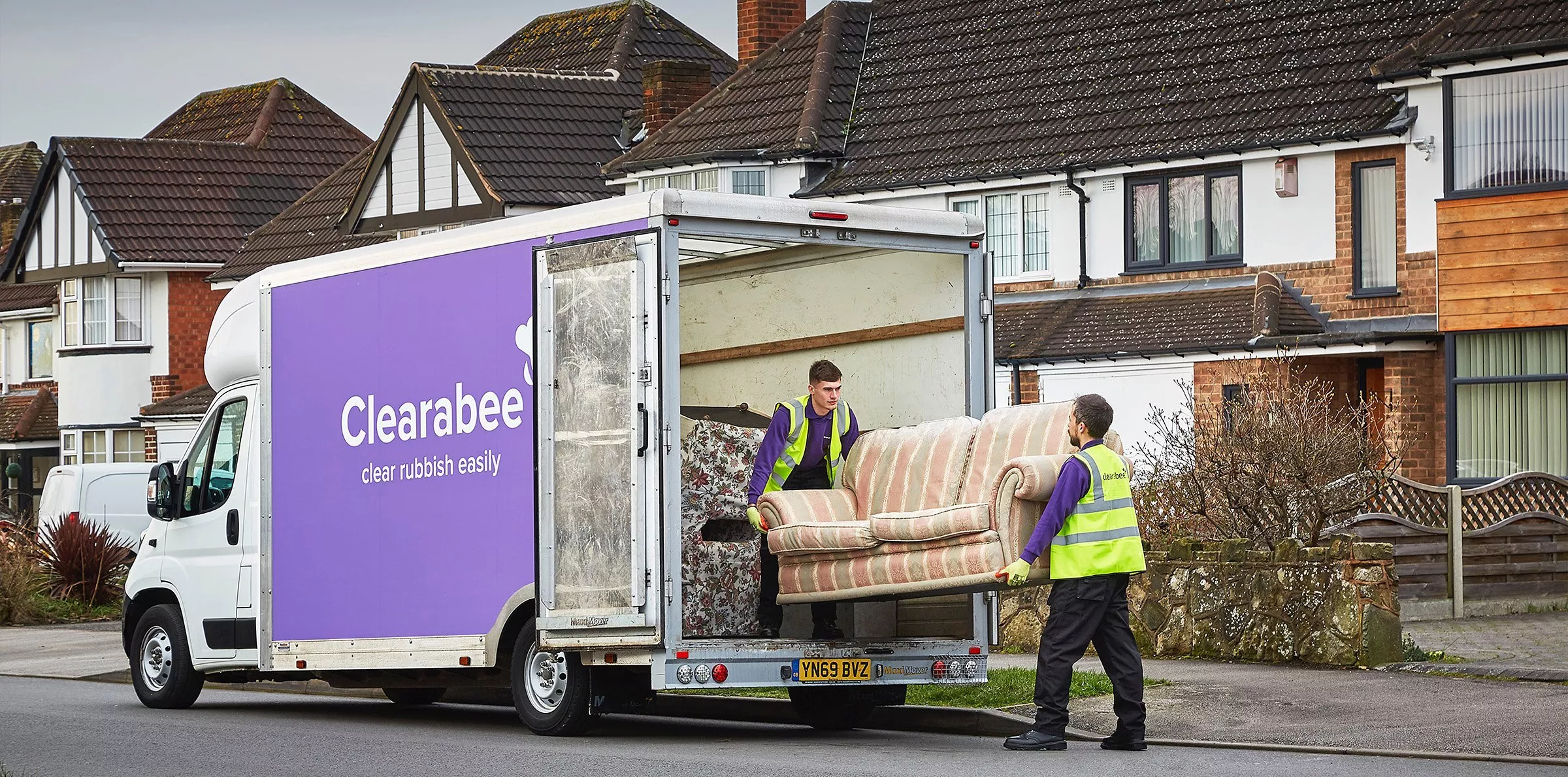 Clearabee rubbish removal crew loading a sofa into a purple clearance van outside suburban UK houses