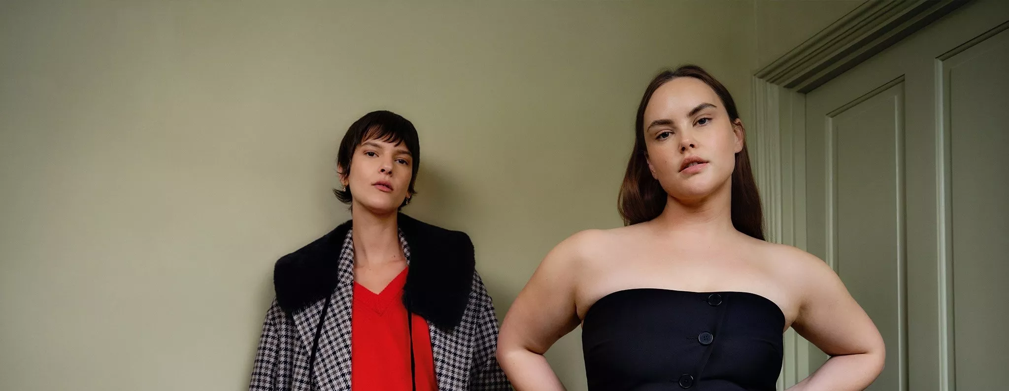 Two women standing, one in a red jumper, checkered coat and blue skirt, the other in black strapless top and black trousers