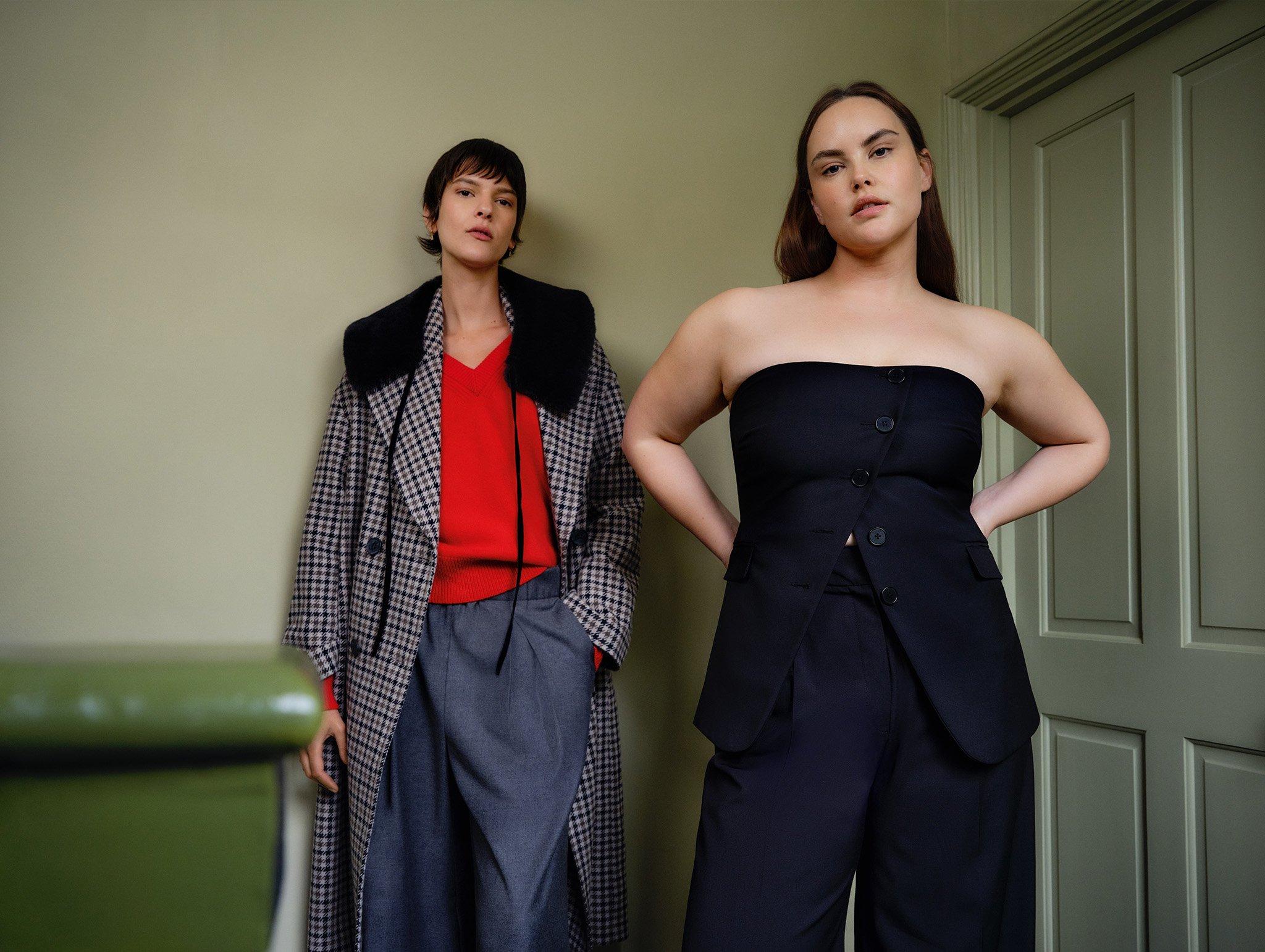 Two women standing, one in a red jumper, checkered coat and blue skirt, the other in black strapless top and black trousers