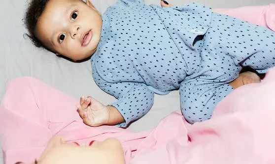 A baby in a blue polka-dot outfit lies on a bed, gazing upwards, with an adult in pink resting beside them.