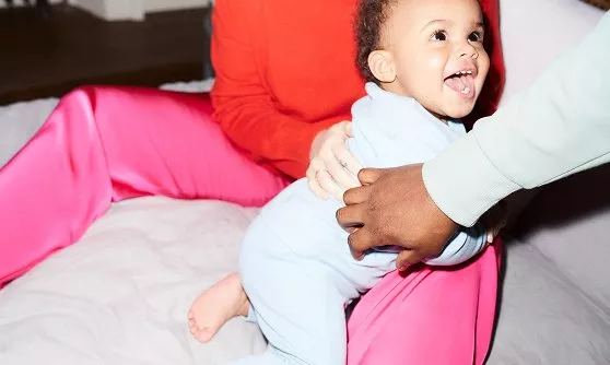 A smiling baby in blue pyjamas is being held by two adults, one wearing bright pink trousers.