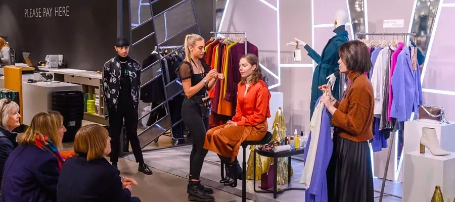 A makeup artist demonstrates on a seated model in a fashion store, surrounded by racks of vibrant clothing.