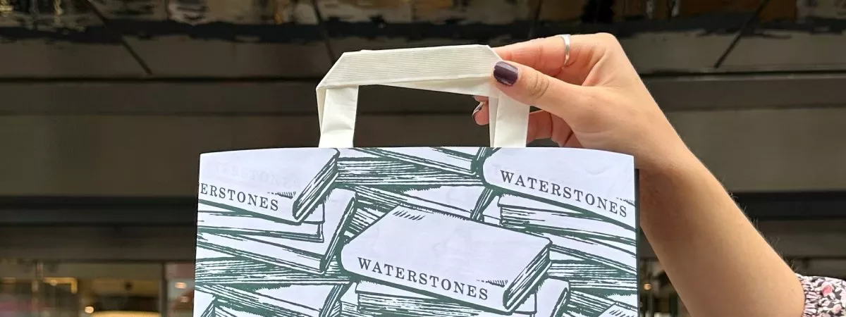 Hand holding a Waterstones bag with book illustrations, against a blurred urban background.