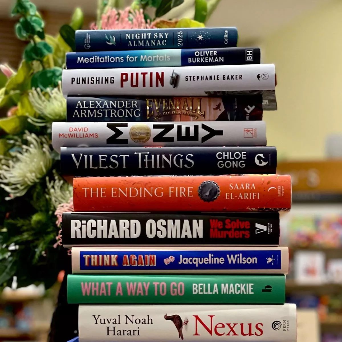 A stack of diverse books on a table, surrounded by greenery. Titles include "Nexus" and "Vilest Things".