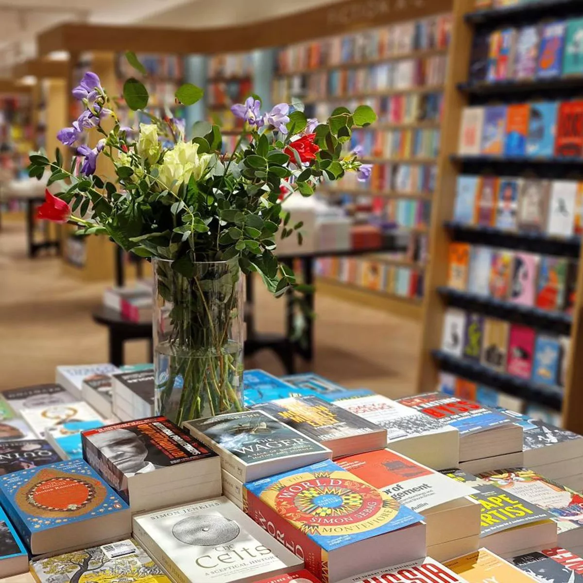 A stack of diverse books on a table, surrounded by greenery. Titles include "Nexus" and "Vilest Things".