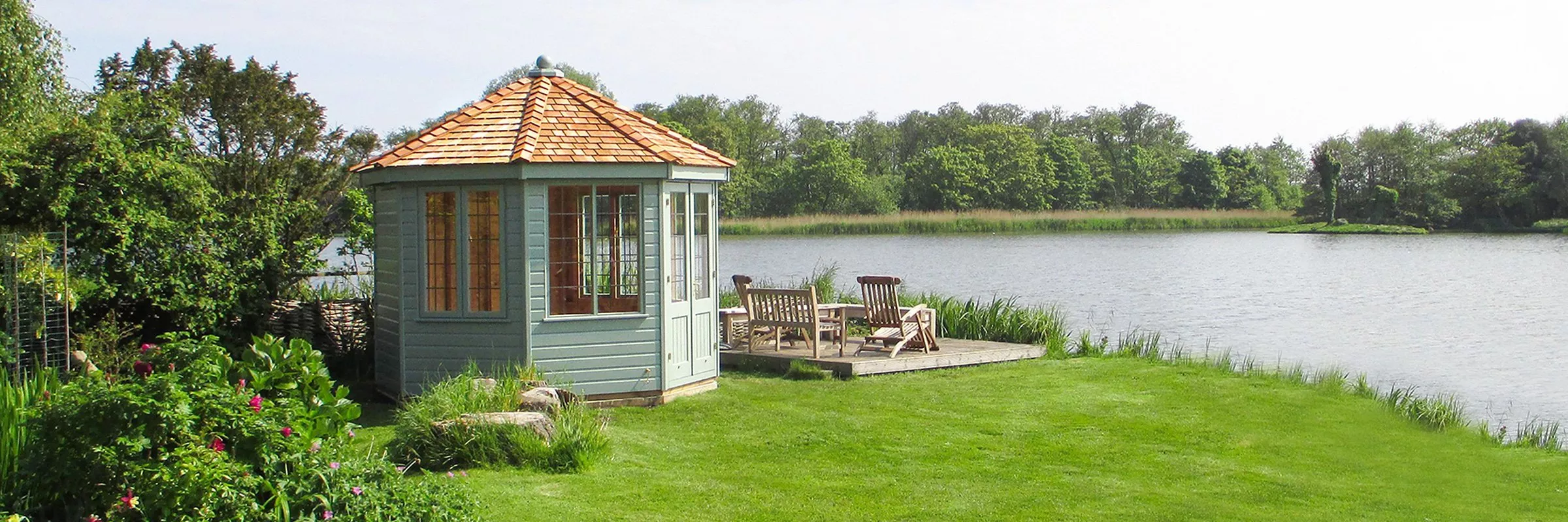 Lakeside garden summerhouse with cedar roof, patio chairs and calm water, surrounded by green trees