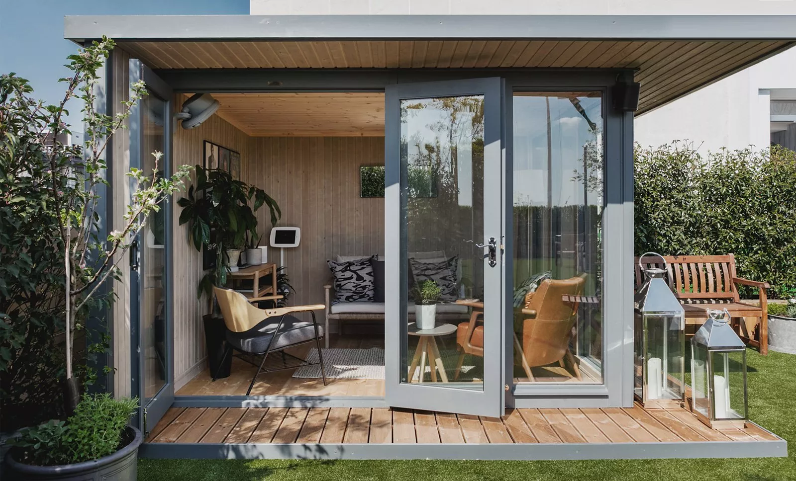 Modern garden office pod with open glass doors, cosy seating, wooden deck and outdoor lanterns in sunlight