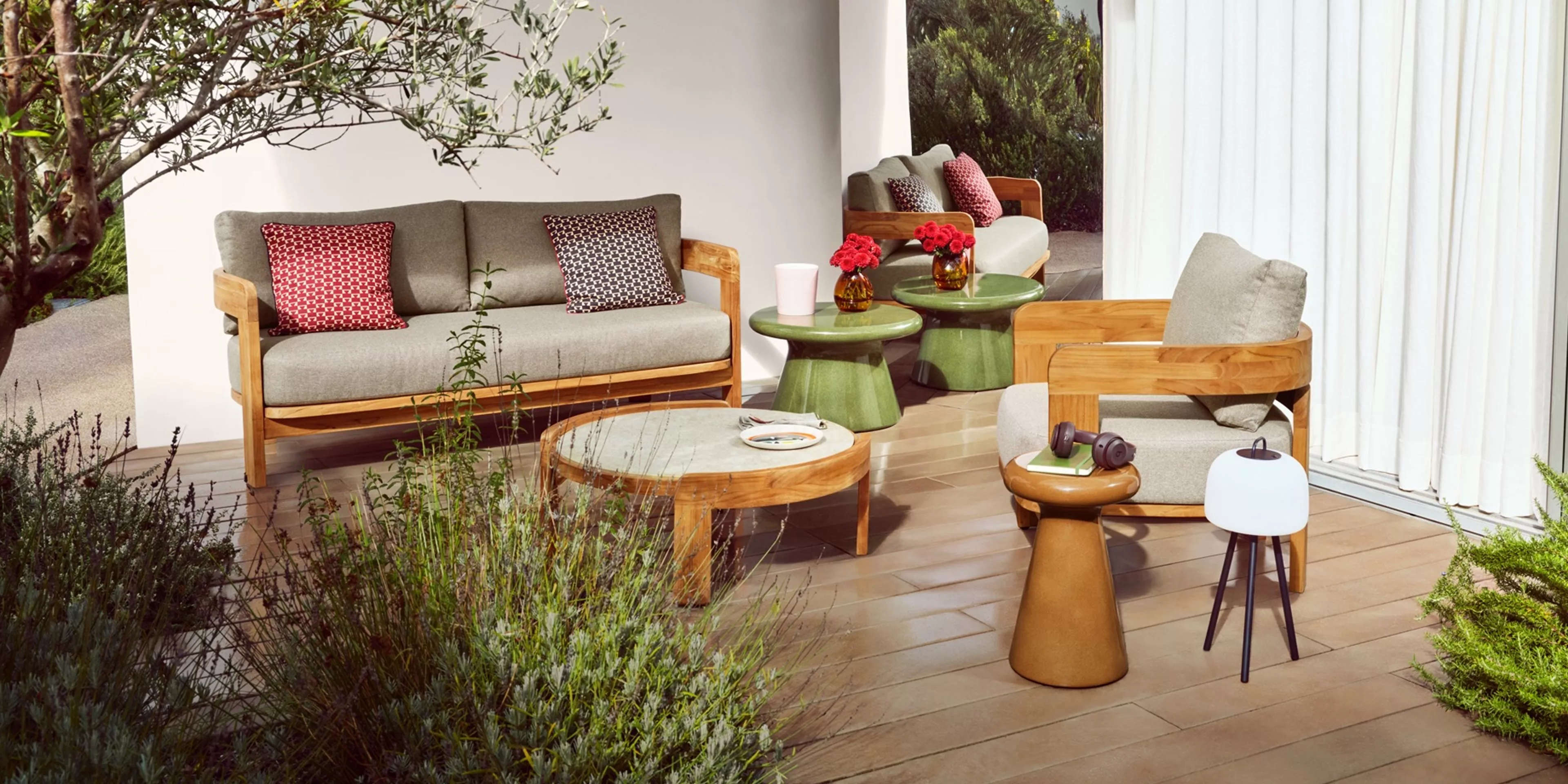 Modern outdoor patio seating with wooden sofa and armchair, green side tables, flowers and garden backdrop