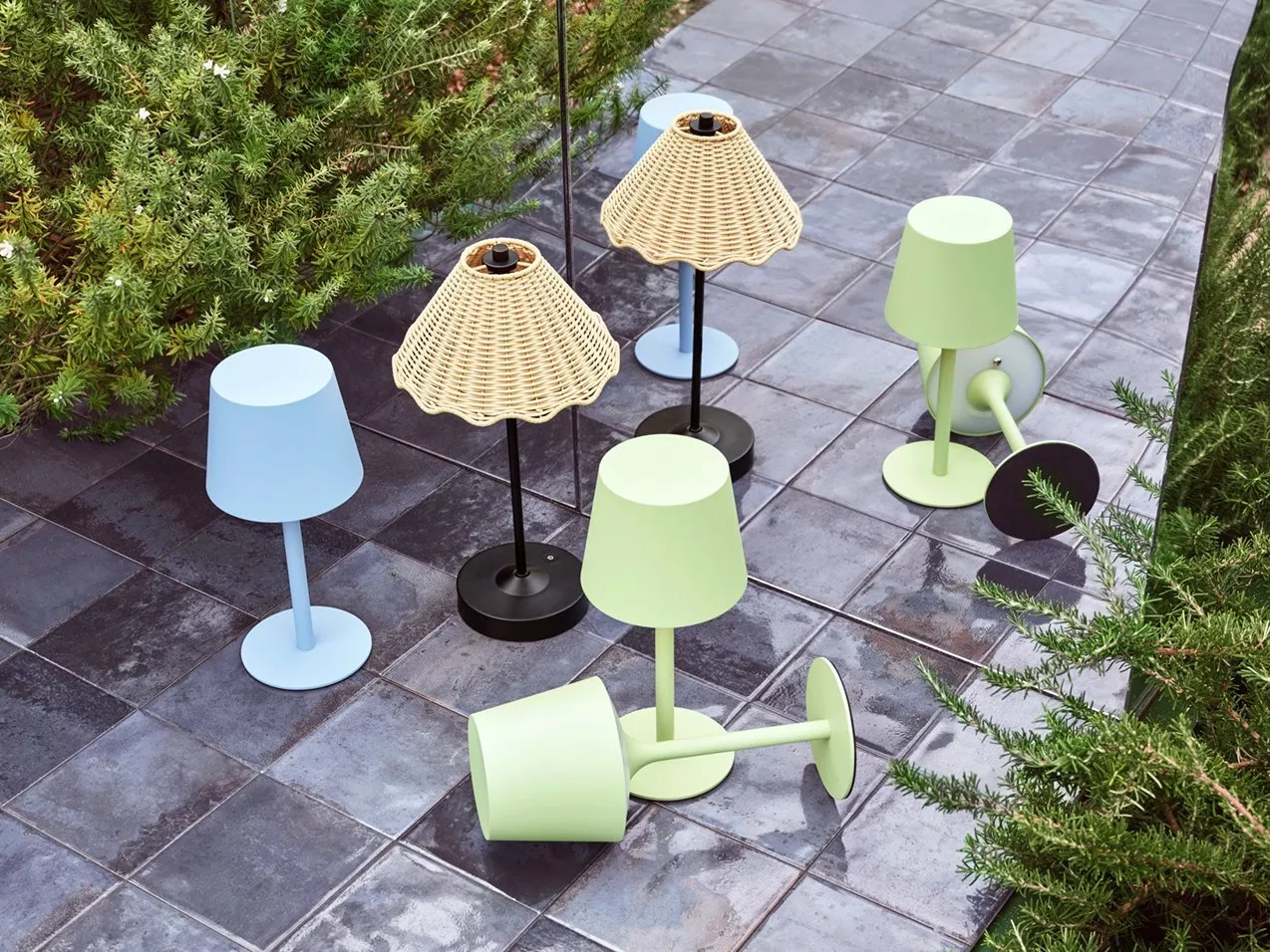 Outdoor patio table lamps in pastel blue and green, plus wicker shades, on wet tiled garden path
