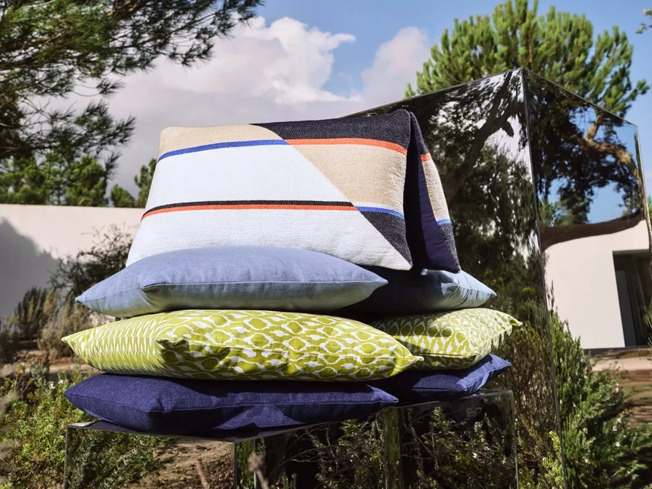 Stacked outdoor cushions with striped throw on mirrored chair in garden setting under blue sky and trees