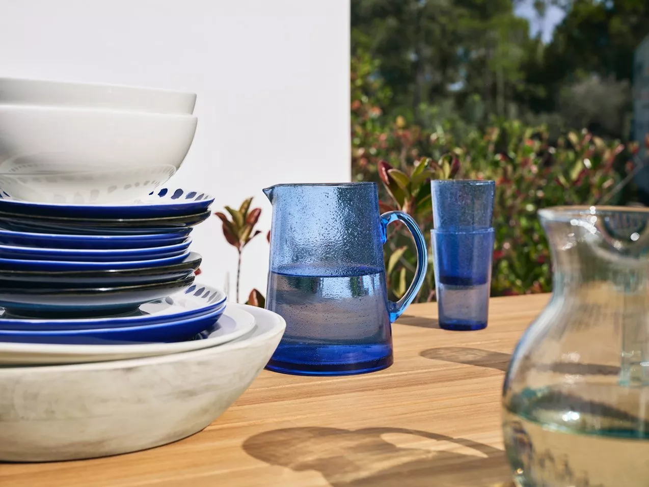 Outdoor dining table with stacked plates and bowls, blue glass water jug and tumblers in sunlight