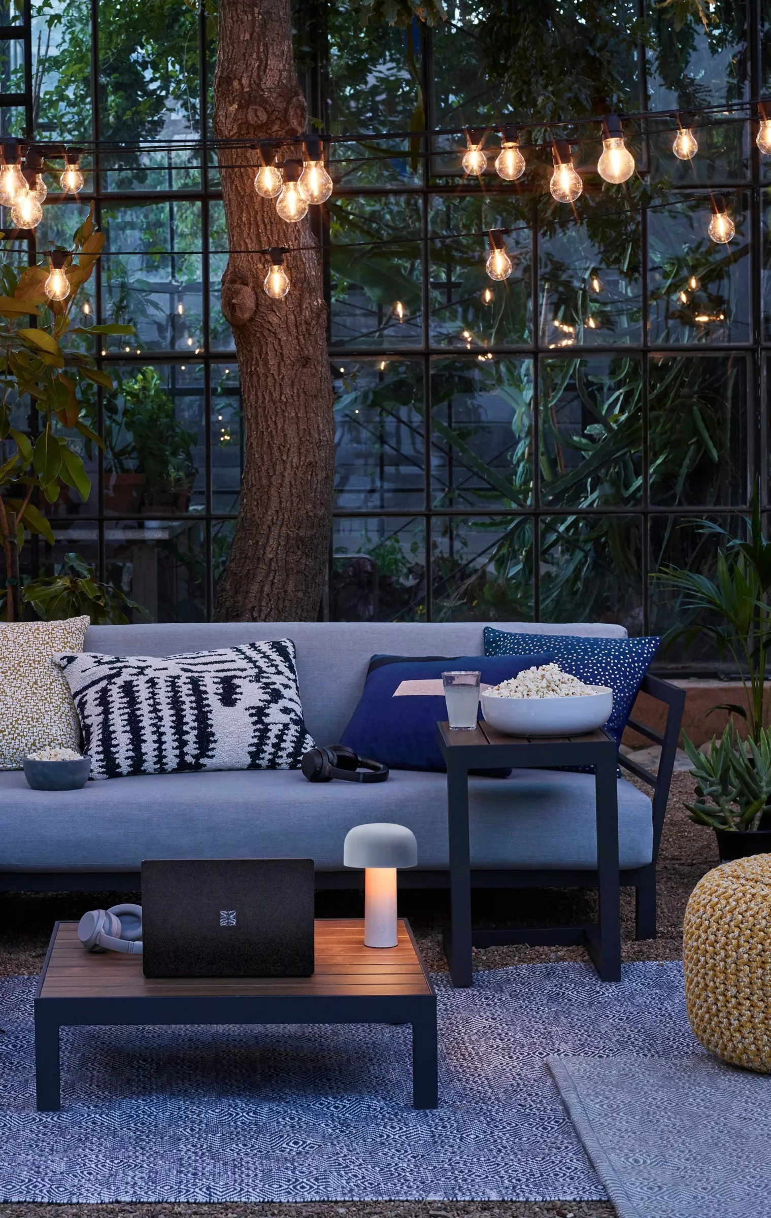 Cosy outdoor lounge with string lights, sofa cushions, popcorn and laptop on patio in glasshouse garden setting