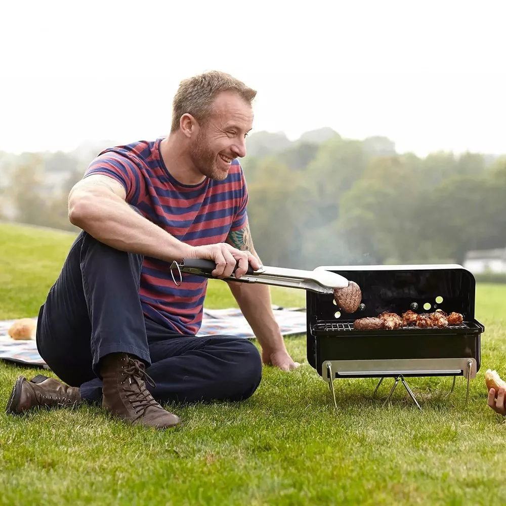 man sitting on the grass next to a weber go-anywhere charcoal bbq flipping a burger