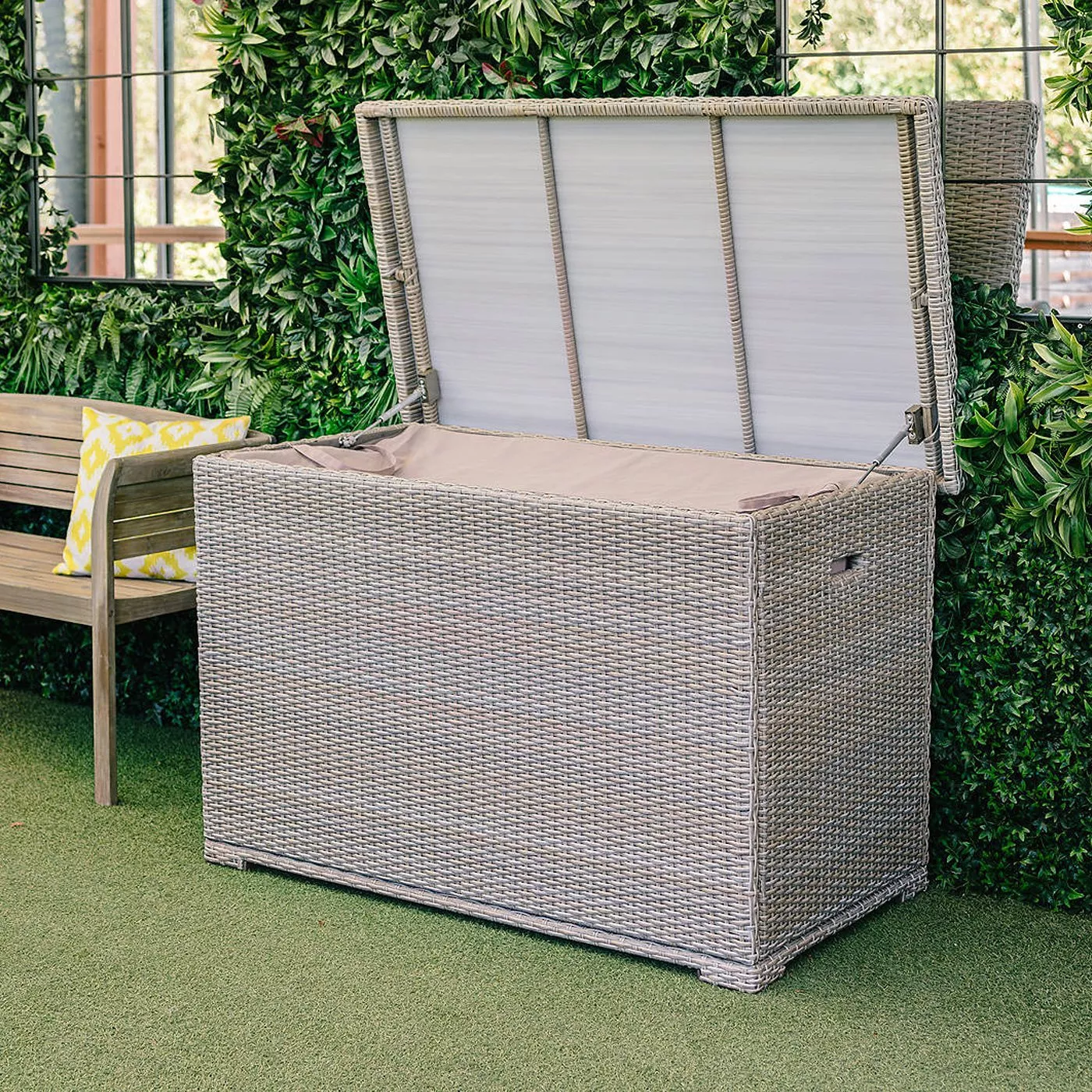 Grey rattan garden storage box with lift-up lid, outdoor cushion chest on patio beside green wall and bench.